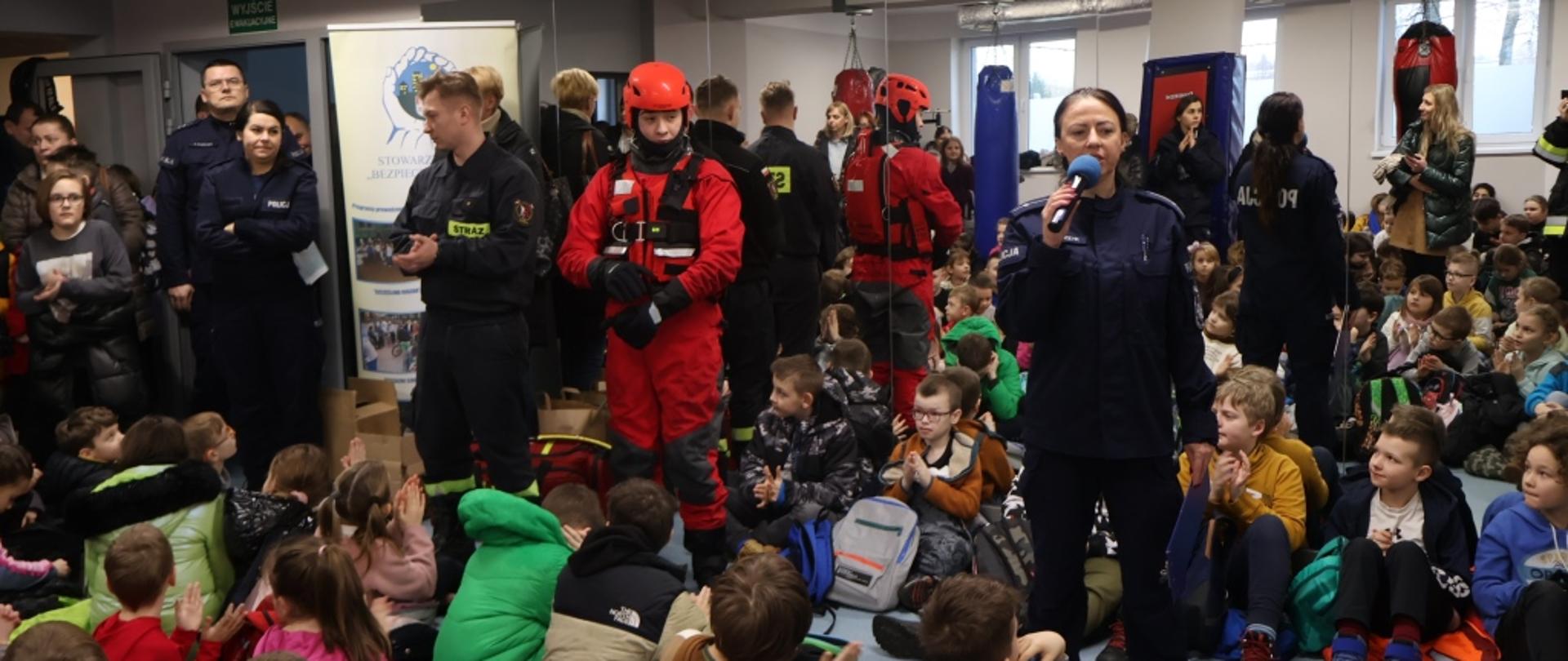 Zdjęcie wykonane w jednej z sali Miejskiego Ośrodka Sportu i Rekreacji w Radomiu na osiedlu Borki. Na zdjęciu widać siedzące na ziemi dzieci. Przed nimi z mikrofonem stoi funkcjonariuszka Policji. Obok niej stoi dwóch funkcjonariuszy Państwowej Straży Pożarnej. Jedne z nich ubrany jest w specjalny kombinezon do prowadzenia działań ratowniczych na lodzie i zamarzniętych akwenach wodnych. W oddali obok widać jeszcze innych stojących funkcjonariuszy Policji oraz rodziców. Ustawieni przed dziećmi funkcjonariusze omawiają zasady bezpiecznego zachowania i zagrożenia jakie mogą wystąpić podczas ferii zimowych. 