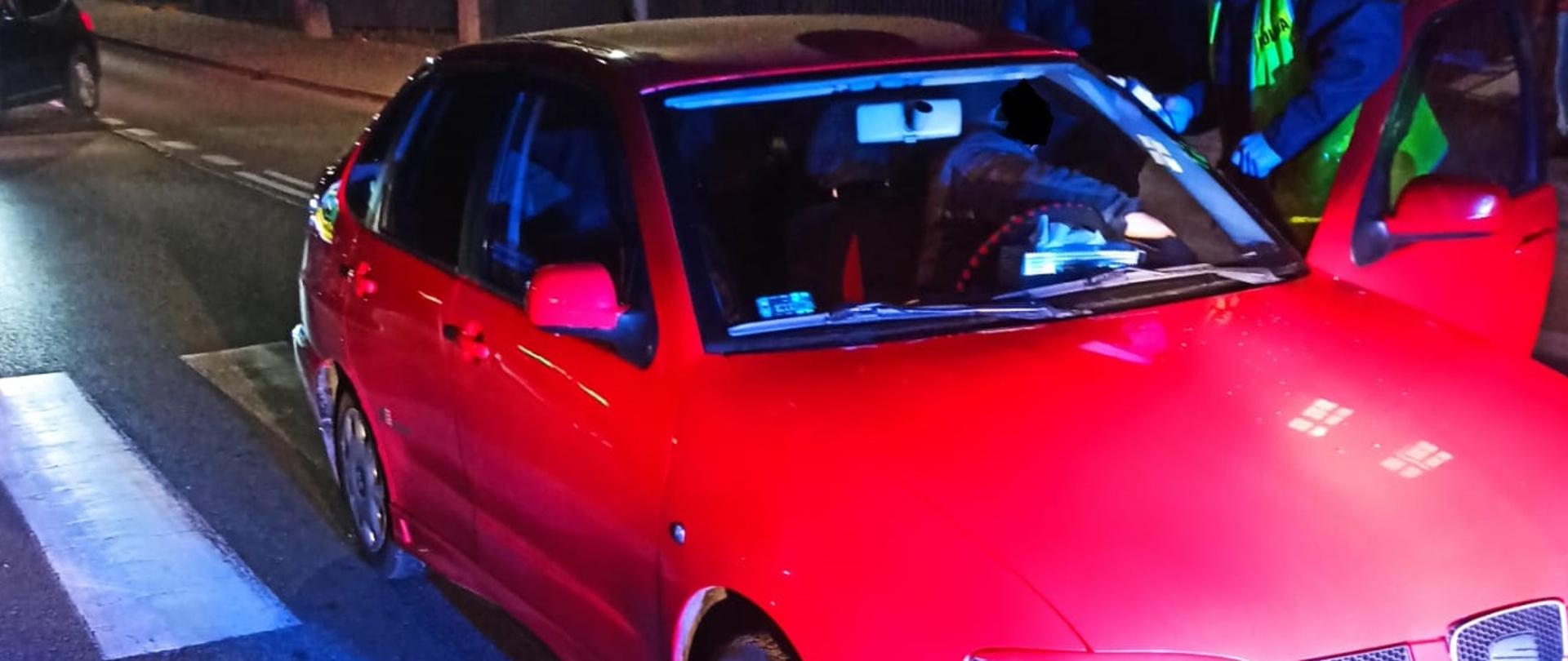 Pora nocna. Czerwony samochód osobowy stoi na pasach. W środku siedzi kierowca ma otwarte drzwi, przy nim stoi policjant. 