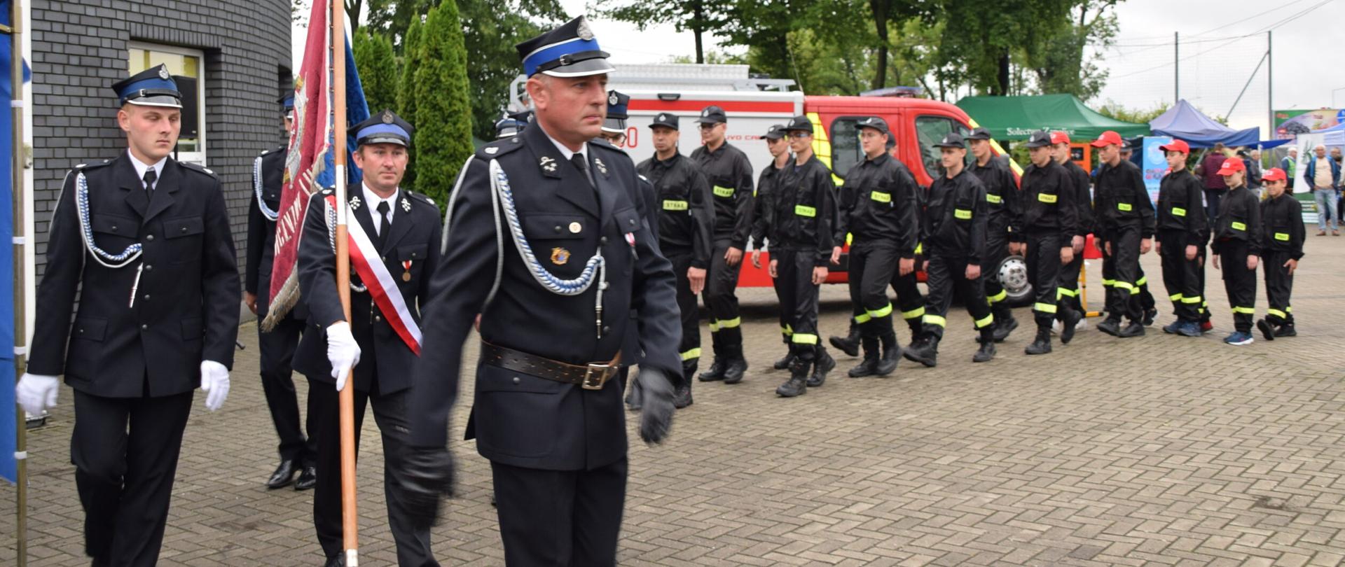 Zdjęcie przedstawia strażaków z OSP Wojsławice podczas przemarszu po placu. Pododdział prowadzony jest przez dowódcę uroczystości, za nim widać poczet sztandarowy oraz pozostałych druhów. 