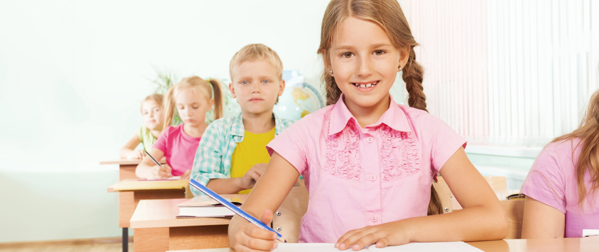 Dzieci siedzące w szkolnych ławkach, jedno za drugim. Na pierwszym planie uśmiechnięta dziewczynka w różowej koszulce z kredką w ręku, za dziewczynką siedzi chłopiec z otwartą książką. Na dalszym planie widać dwie dziewczynki siedzące jedna za drugą. 