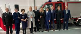 Zdjęcie grupowe na tle wozu strażackiego, w jednym rzędzie stoją mężczyźni ubrani w mundury wyjściowe PSP, OSP oraz osoby w strojach cywlinych