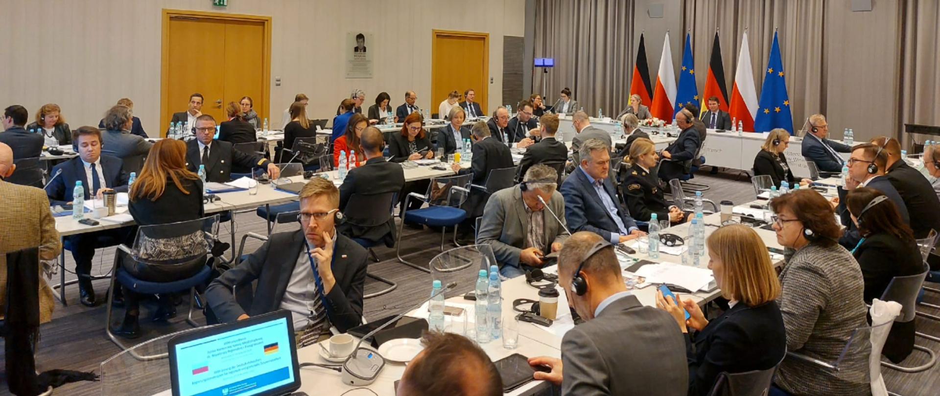 Widok ogólny na sale w której odbywa się posiedzenie Polsko-Niemieckiej Komisji Miedzyrządowej ds. Współpracy Regionalnej i Przygranicznej. Osoby siedzące przy stołach, w tle stolik prezydialny za którym widoczne są flagi Polski, Niemiec i Unii Europejskiej