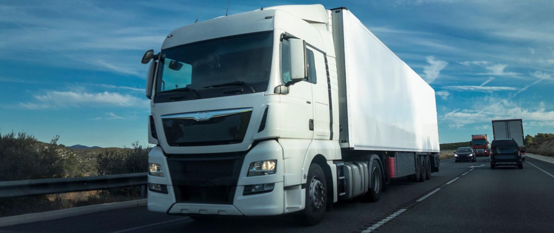 na zdjęciu widnieje duży, biały samochód ciężarowy. W tle widać samochody osobowe, które za nim jadą oraz inne samochody ciężarowe.