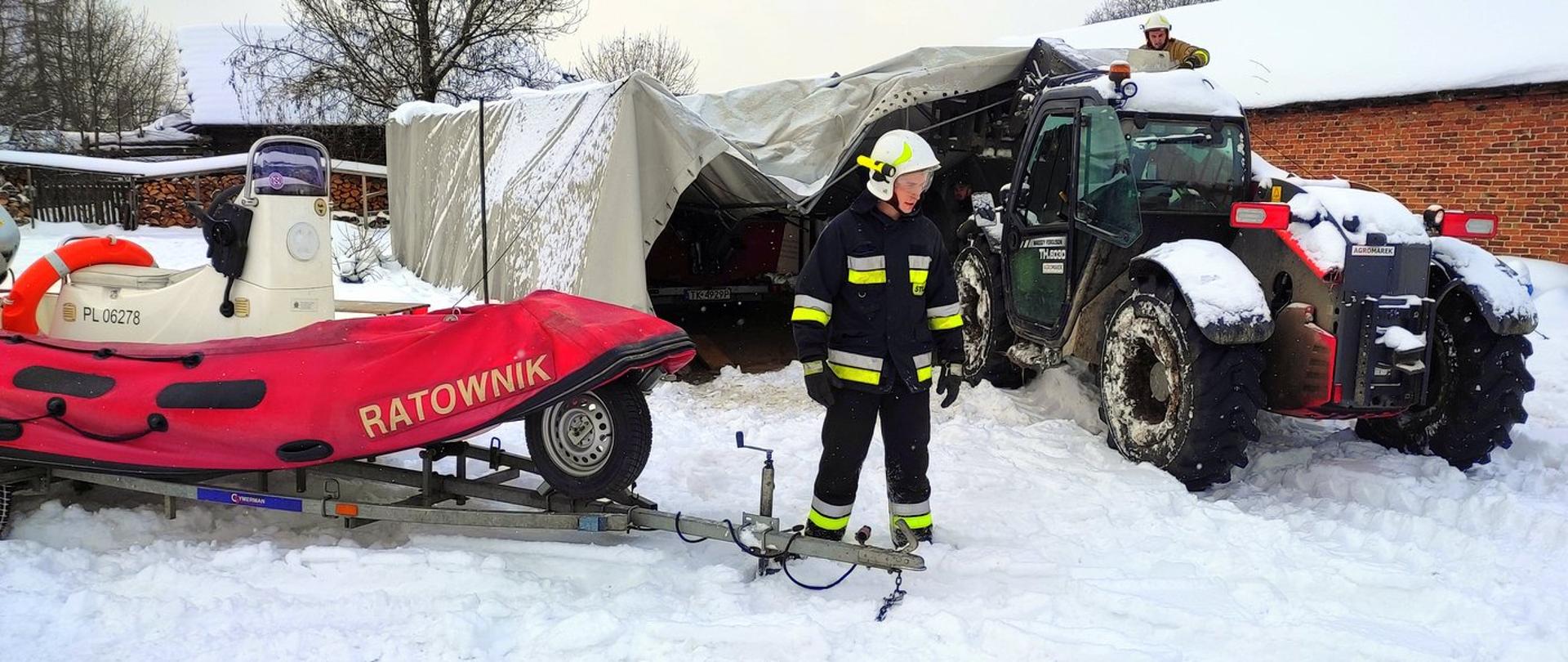 Zdjęcie wykonane na zewnątrz. Po lewej stronie, na przyczepie motorowa Łódź ratownicza z napisem "ratownik". Obok przyczepy stoi strażak. W tle widać namiot, który zawalił się pod naporem śniegu. 