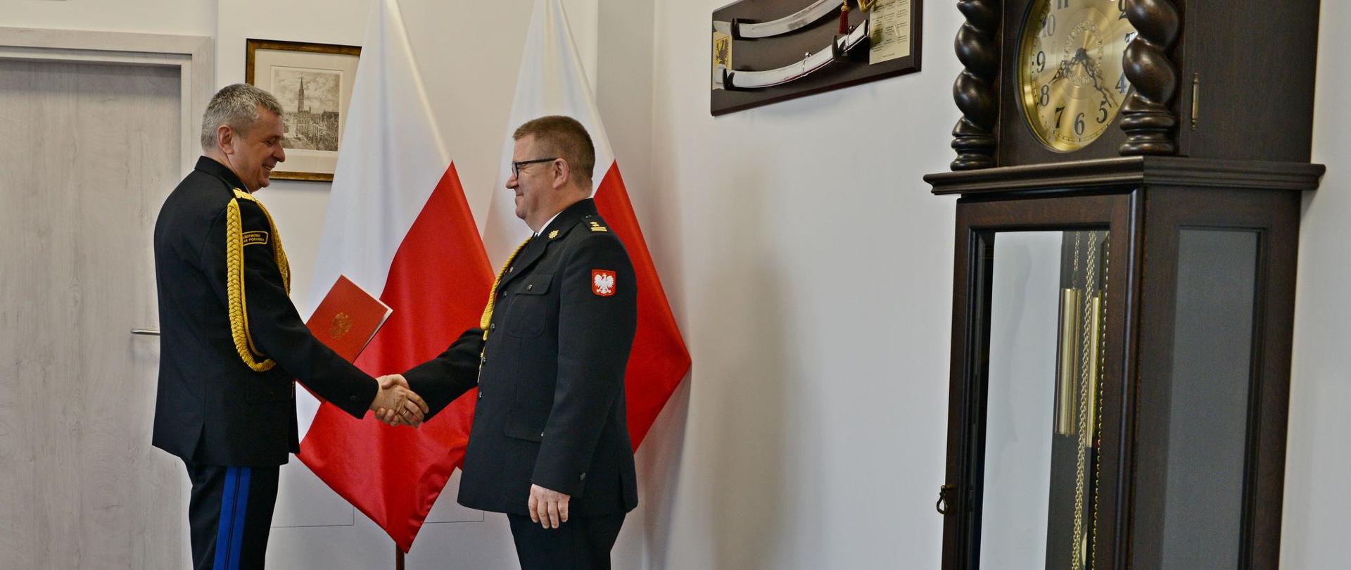 Pomorski komendant wojewódzki Państwowej Straży Pożarnej trzyma czerwoną teczkę jednocześnie trzyma dłoń strażaka oboje mężczyźni ubrani w umundurowanie wyjściowe ze sznurem.