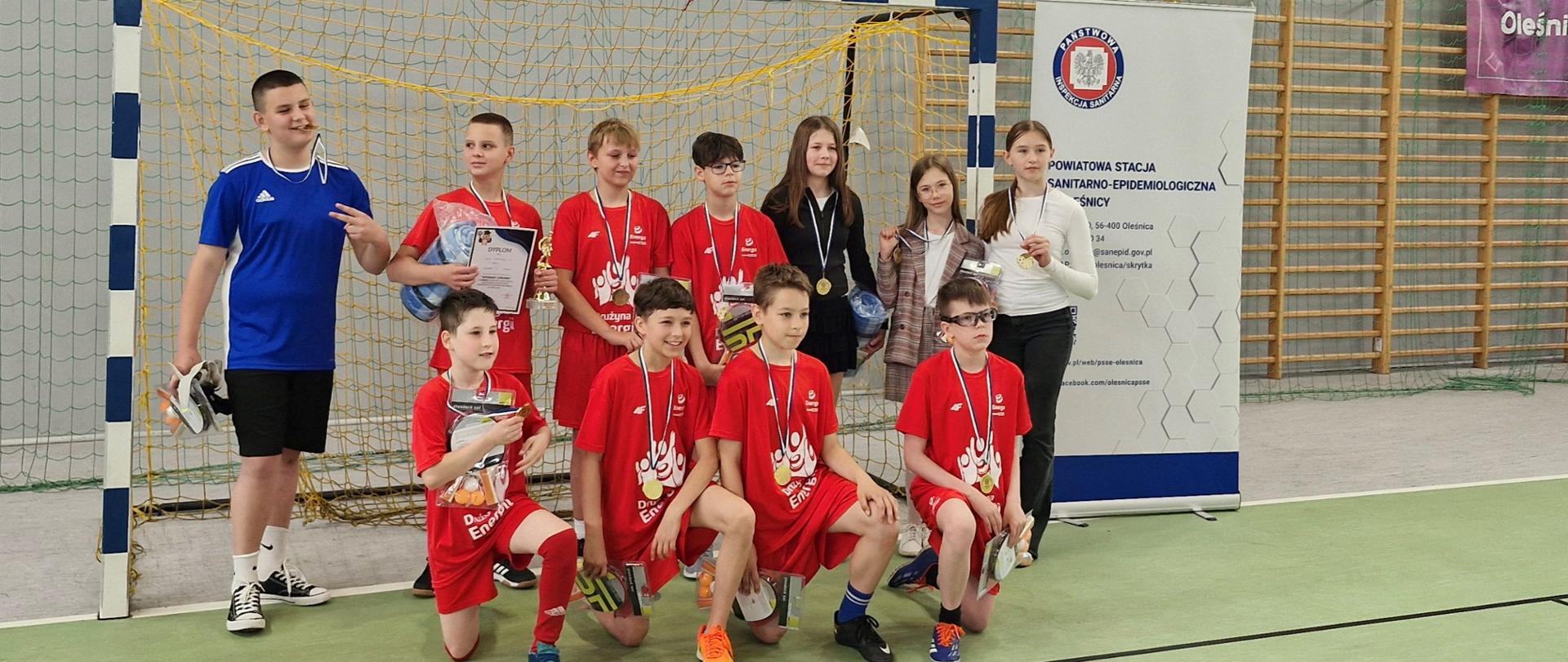 Drużyna piłkarska pozująca do zdjęcia stoi w bramce. W tle baner Państwowej Inspekcji Sanitarnej.