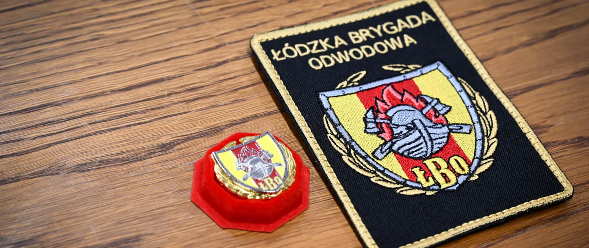 Zbliżenie na czarną, haftowaną naszywkę z napisem „Łódzka Brygada Odwodowa ŁBO” oraz pamiątkową odznakę na czerwonym aksamicie.