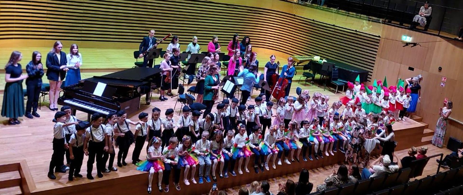 Około 90 dzieci siedzi i stoi w dwóch rzędach na estradzie, za nimi stoją muzycy orkiestry.
