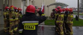 Zdjęcie przedstawia uroczystą zmianę służby przed bramami JRG, strażacy ubrani w umundurowanie specjalne oddają honor, poczet flagowy wciąga flagę RP na maszt.