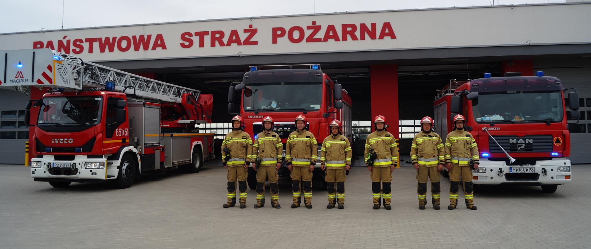 Zdjęcie przedstawia siedmiu strażaków stojących przed pojazdami pożarniczymi
