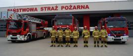 Zdjęcie przedstawia siedmiu strażaków stojących przed wozami pożarniczymi
