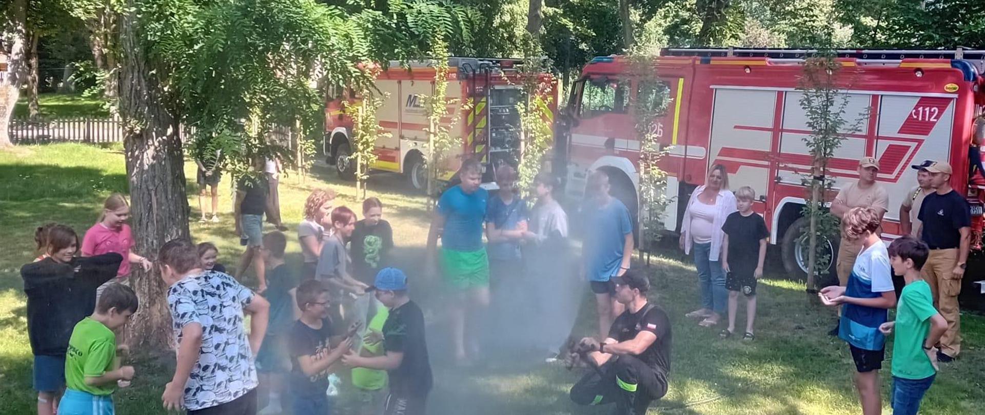 W parku ustawiane dwa samochody strażackie. Dzieci bawią się na trawie. Słoniecznie, zielone drzewa.