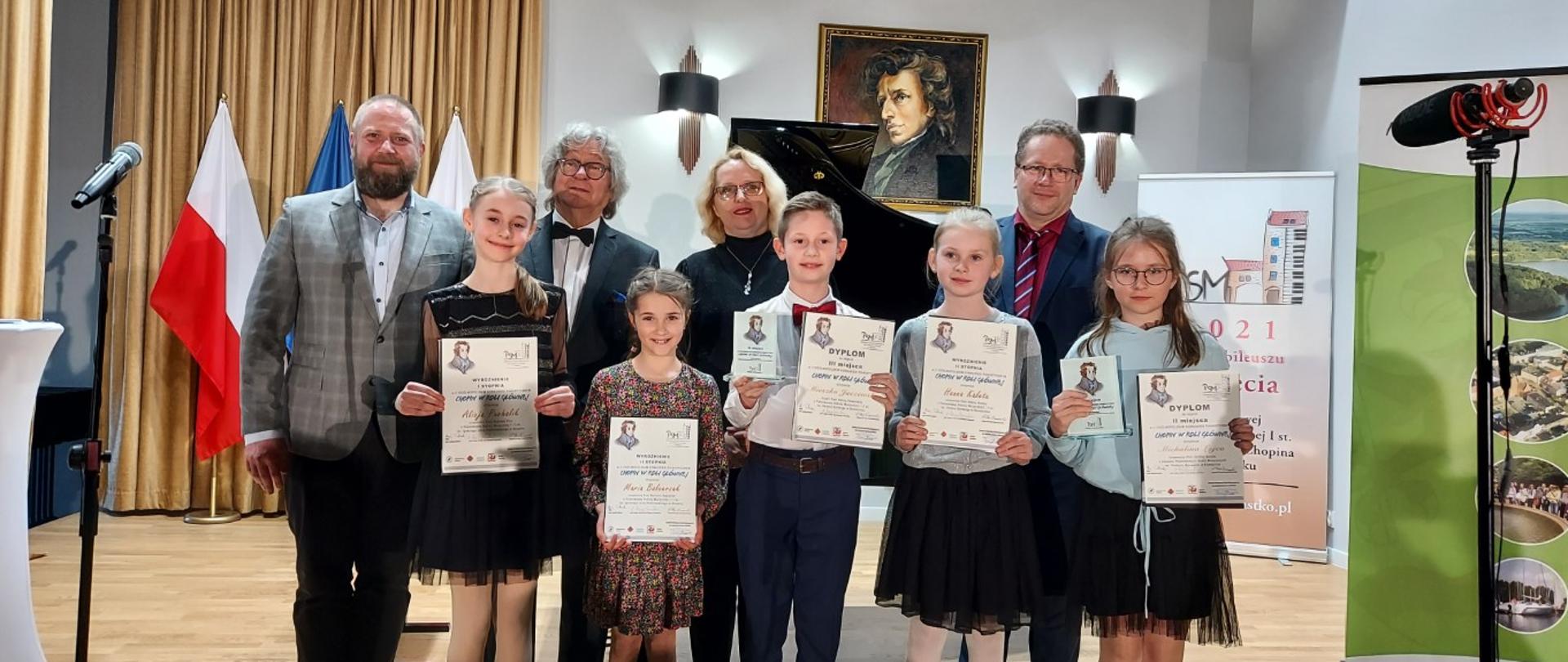 Zdjęcie kolorowe. Na tle fortepianu i portretu Fryderyka Chopina w sali kameralnej szkoły muzycznej w Miastku stoją cztery osoby dorosłe: dyrektor szkoły muzycznej w Miastku i członkowie Jury. Przed nimi stoją laureaci najmłodszej grupy: cztery dziewczynki i jeden chłopiec, dzieci są uśmiechnięte, bo trzymają w rękach dyplomy laureatów. 