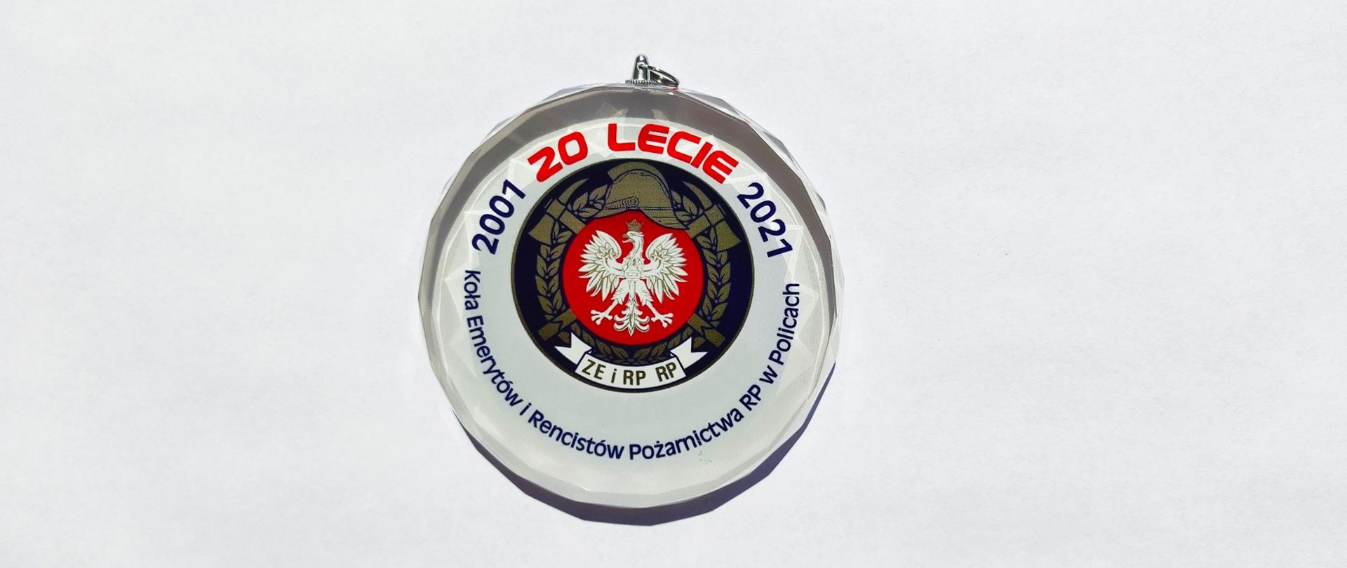 Na zdjęciu okolicznościowy biały medal ze szkła z napisami na obwodzie. Czerwoną czcionką: 20 lecie. Granatową napis: Koła Emerytów i Rencistów Pożarnictwa RP w Policach. W środku logo na granatowym tle: biały orzeł w koronie na czerwonym polu, nad nim złote: historyczny hełm strażacki, skrzyżowane toporki, poniżej biała szarfa z napisem ZE i RP RP. Całość środkowego pola okalają złote liście laurowe.