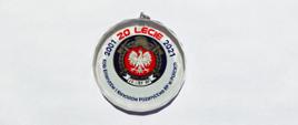 Na zdjęciu okolicznościowy biały medal ze szkła z napisami na obwodzie. Czerwoną czcionką: 20 lecie. Granatową napis: Koła Emerytów i Rencistów Pożarnictwa RP w Policach. W środku logo na granatowym tle: biały orzeł w koronie na czerwonym polu, nad nim złote: historyczny hełm strażacki, skrzyżowane toporki, poniżej biała szarfa z napisem ZE i RP RP. Całość środkowego pola okalają złote liście laurowe.