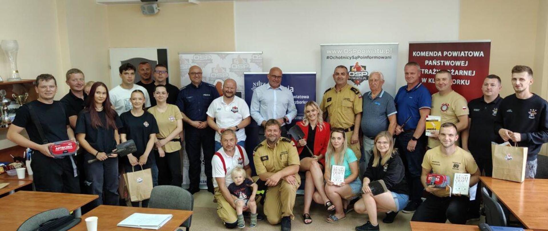 Zdjęcie przedstawia grupę ludzi pozujących do wspólnego zdjęcia w pomieszczeniu biurowym. W tle znajdują się trzy banery z logo organizacji strażackiej OSP oraz informacjami w języku polskim. Na pierwszym planie po lewej stronie stoi młody mężczyzna trzymający apteczkę pierwszej pomocy. Obok niego stoi grupa młodych ludzi, w tym dwie kobiety i mężczyzna, którzy także trzymają różne przedmioty związane z bezpieczeństwem. W środku grupy, przed banerami, stoi kilku mężczyzn ubranych w mundury strażackie i policyjne, w tym starszy mężczyzna z małym dzieckiem na rękach. Wśród nich są również dwie kobiety siedzące na krzesłach, trzymające książki. Po prawej stronie grupy znajdują się kolejni mężczyźni w mundurach, a wśród nich także młody mężczyzna trzymający torbę. Pomieszczenie jest jasne, z oknami po lewej stronie, a na biurkach widać różne dokumenty i kubki.