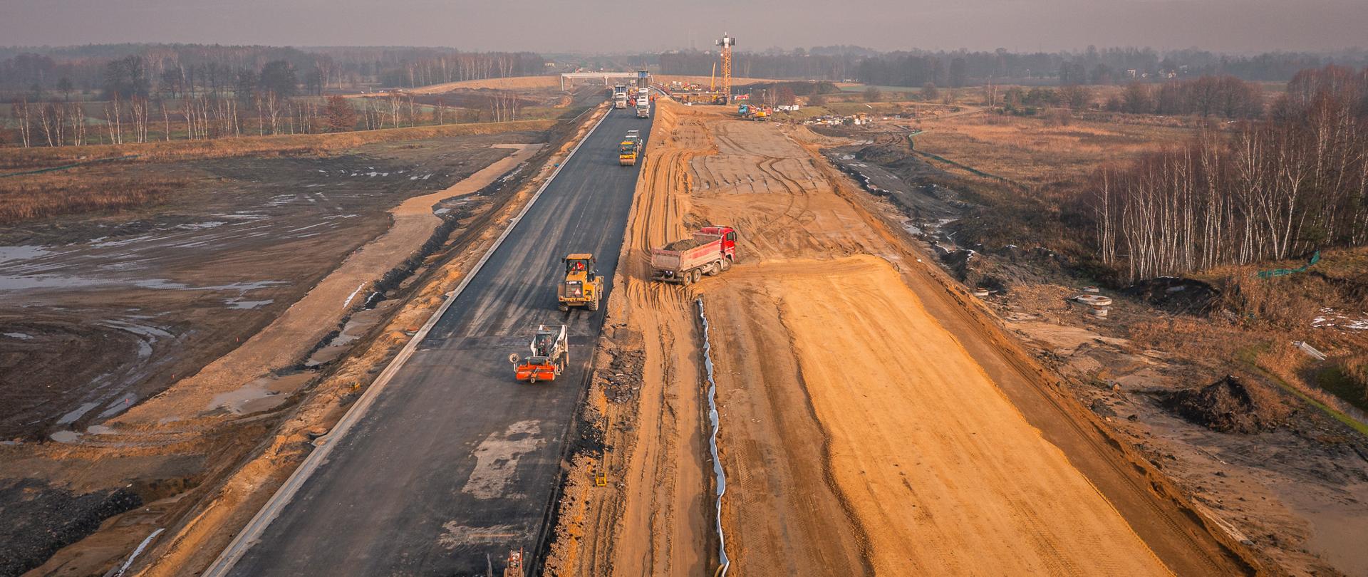 Widok z góry na plac budowy drogi. Po lewej stronie widać fragment ciemnej, wstępnie ułożonej nawierzchni asfaltowej, na której pracują maszyny drogowe. Po prawej znajdują się szerokie pasy jasnego, świeżo wyrównanego nasypu ziemnego. Na całej długości budowy ustawiono kilka pojazdów i maszyn budowlanych. Teren wokół jest otwarty, z polami i zadrzewieniami w tle. 