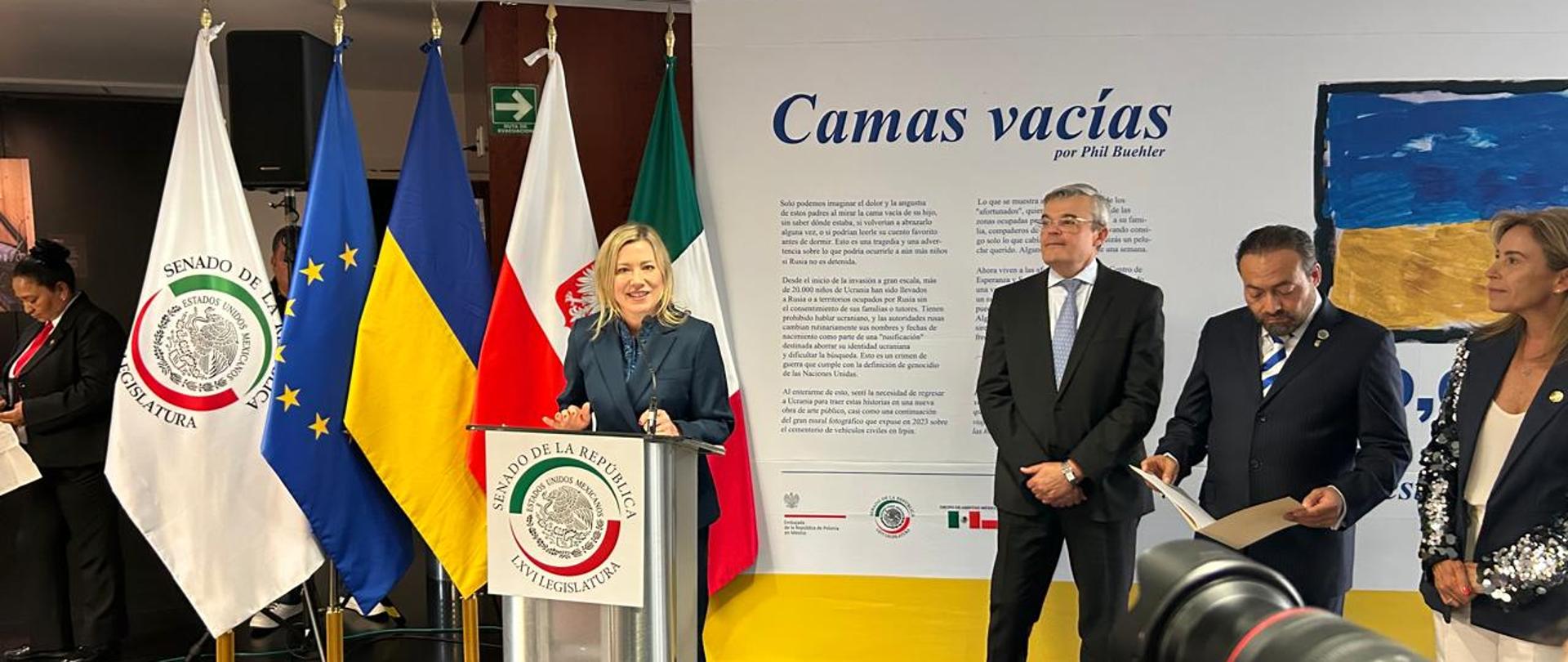 La Embajadora Agnieszka Frydrychowicz-Tekieli pronuncia un discurso durante la inauguración de la exposición. Al fondo se observan las banderas de México, Polonia, Ucrania, la Unión Europea y el Senado de México, así como el panel introductorio de la exposición “Camas Vacías”.