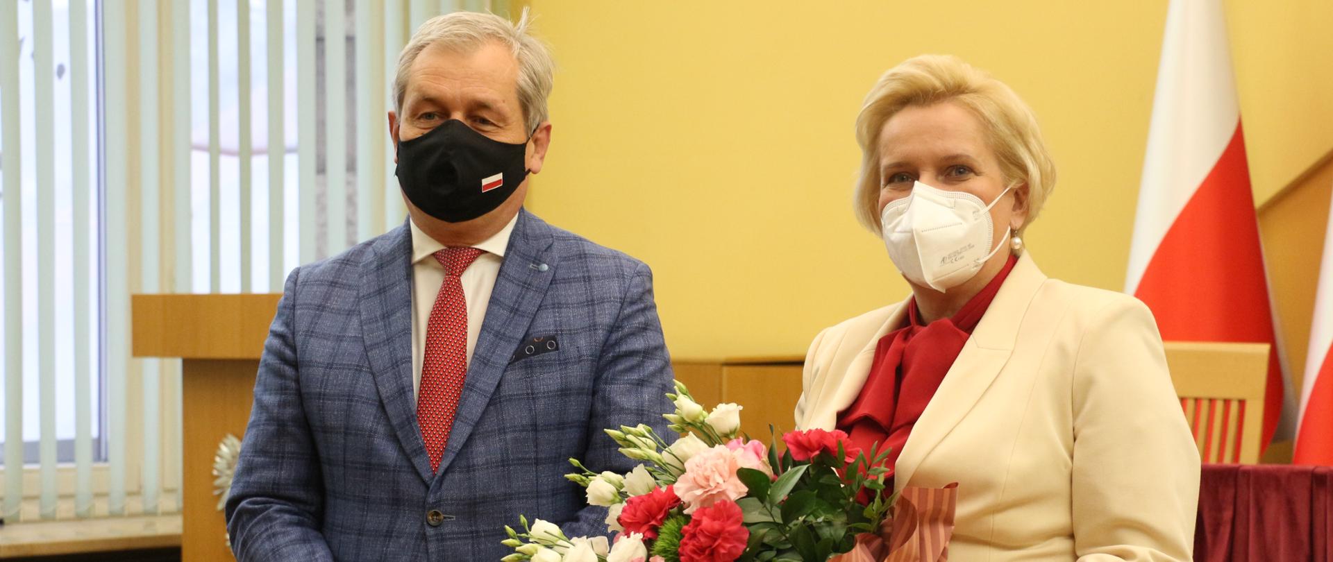 Na zdjęciu wojewoda i wicewojewoda opolski. Po lewej stronie wojewoda opolski, pozujący do zdjęcia. Po prawej wicewojewoda opolski trzymająca bukiet kwiatów.