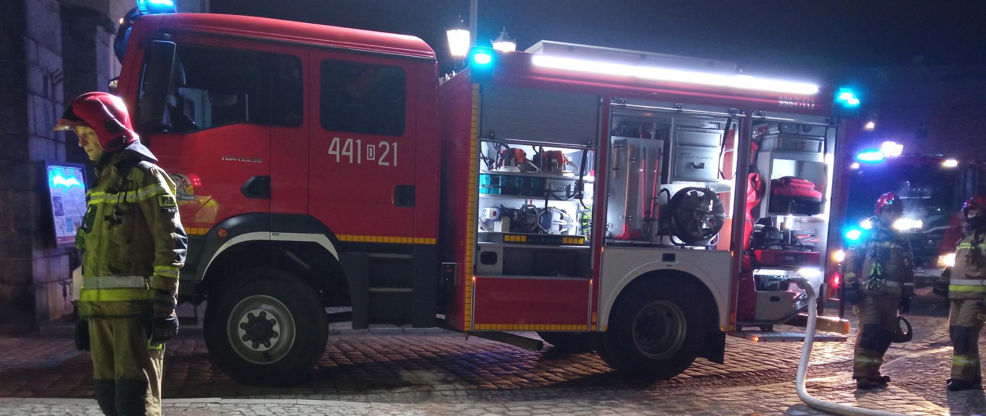 Obraz przedstawia samochód pożarniczy z włączonymi sygnałami świetlnymi. Obok strażacy w umundurowaniu specjalnym. Pora nocna.