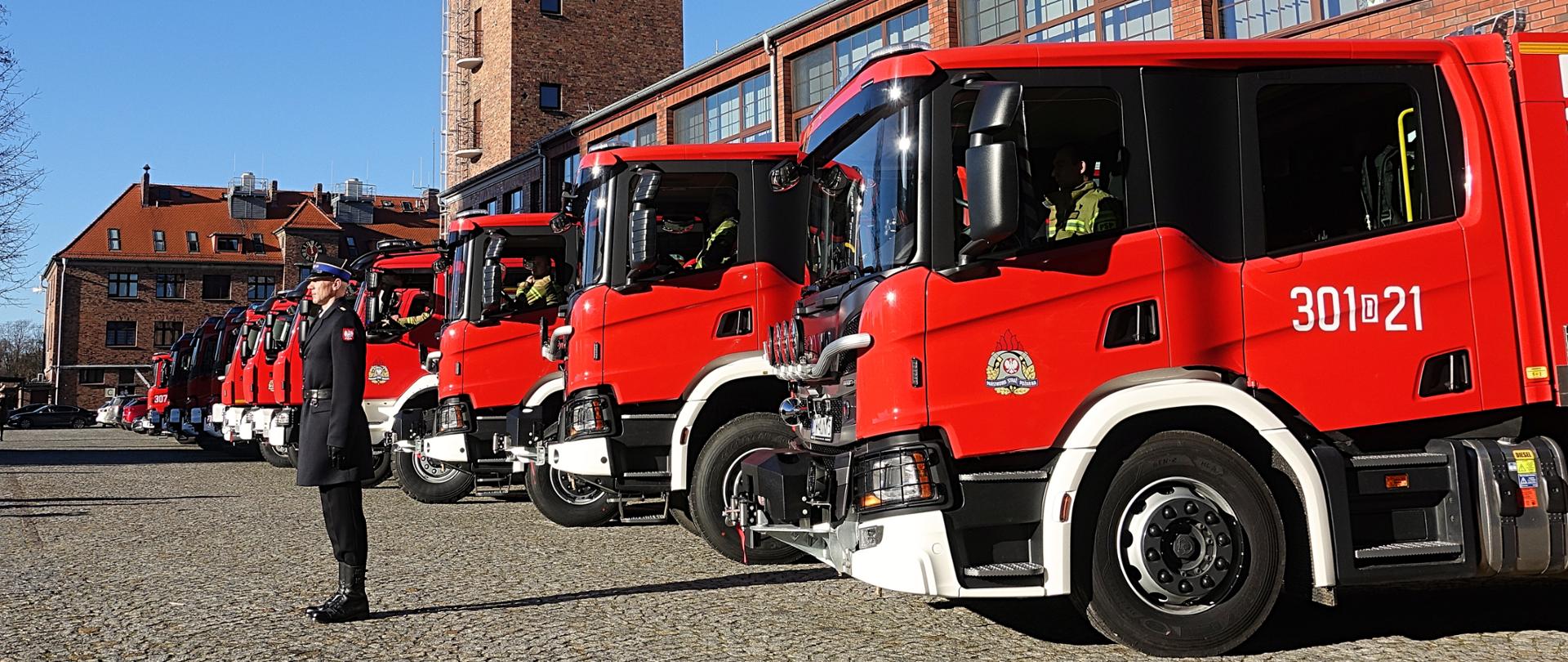Samochody pożarnicze ustawione w rzędzie przed budynkiem strażnicy. Przed samochodami stojący oficer w umundurowaniu galowym. 