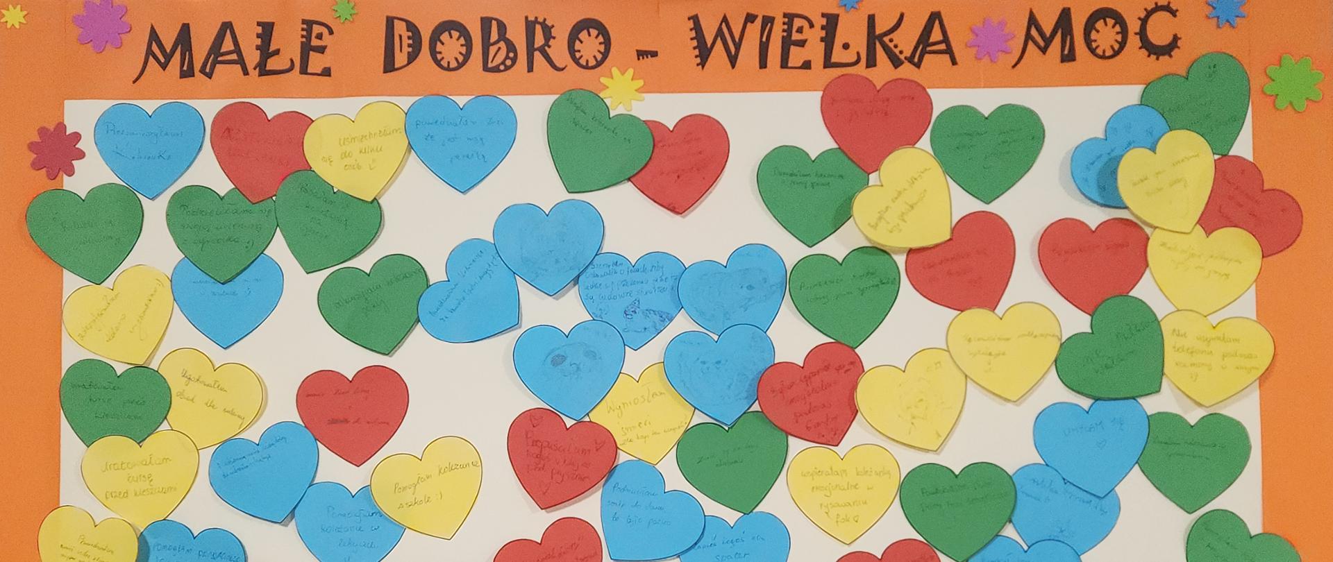 Biała tablica naklejona na pomarańczowy karton na górze napis "MAŁE DOBRO - WIELKA MOC" i kolorowe gwiazdki. Na białym kartonie jest przyklejonych kilkadziesiąt kolorowych - czerwone, zielone, niebieskie i żółte - serduszek z napisami