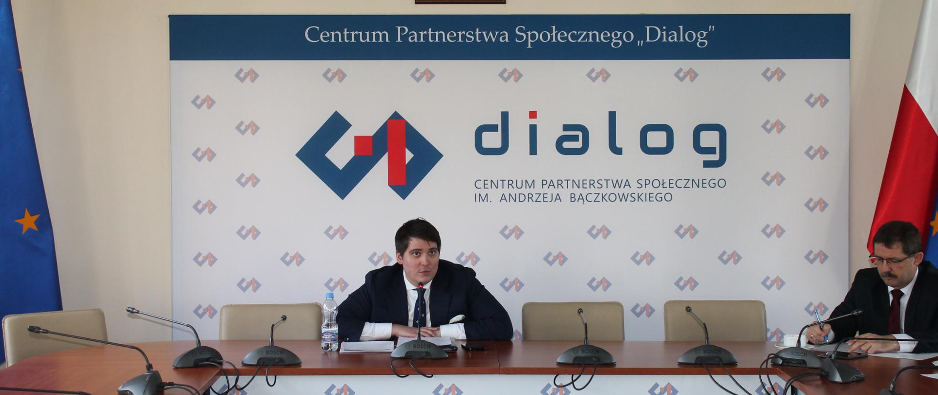 Zdjęcie przedstawia stół prezydialny za którym siedzą: Pan Jakub Bińkowski, Przewodniczący Zespołu oraz Pan Paweł Zgórzyński, Podsekretarz Stanu w Ministerstwie Rodziny, Pracy i Polityki Społecznej. W tle znajduje się baner z napisem Centrum Partnerstwa Społecznego im. Andrzeja Bączkowskiego. Nad banerem znajduje się Godło Polski. Po prawej stronie stoi flaga Polski, po lewej - flaga Unii Europejskiej