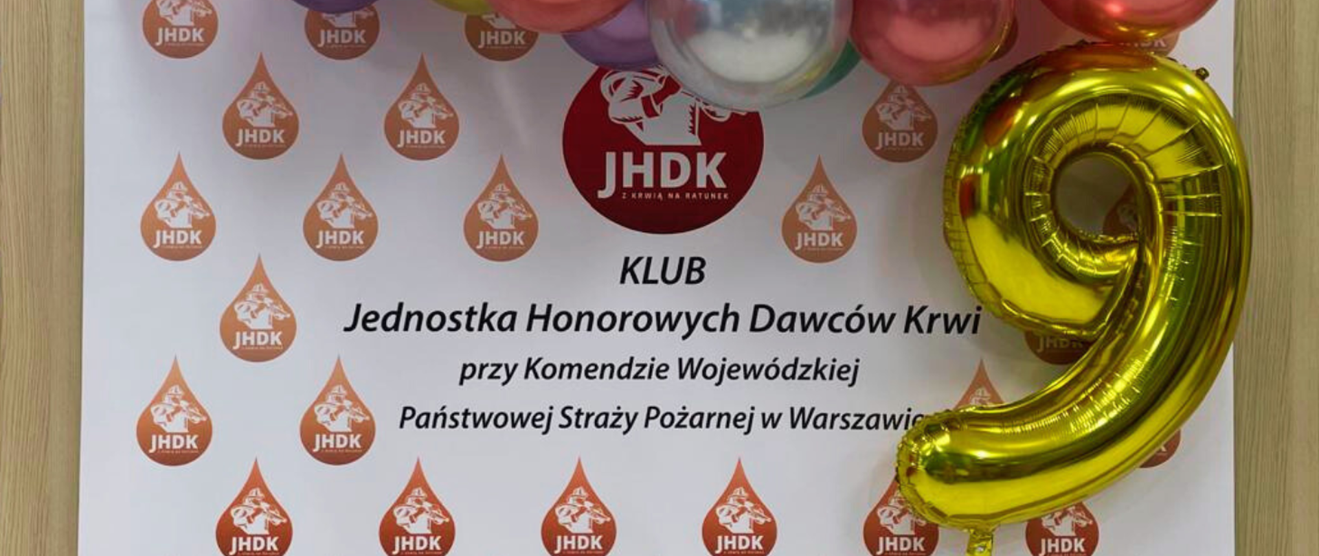 Na zdjęciu biało-czerwony roll-up z napisem „KLUB Jednostka Honorowych Dawców Krwi przy Komendzie Wojewódzkiej Państwowej Straży Pożarnej w Warszawie” oraz duży złoty balon w kształcie cyfry 9. 
