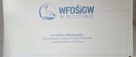 Tablica informacyjna z logo Wojewódzkiego Funduszu Ochrony Środowiska i Gospodarki Wodnej w Olsztynie. Na tablicy znajduje się niebieski napis „WFOŚiGW w Olsztynie” oraz grafika przedstawiająca sylwetkę ptaka nad falami w okręgu. Poniżej widnieje tekst: „Korzystamy z dofinansowania Wojewódzkiego Funduszu Ochrony Środowiska i Gospodarki Wodnej w Olsztynie”. Tło jest jasnoszare, a tablica zamocowana jest na ścianie za pomocą metalowych uchwytów w rogach.