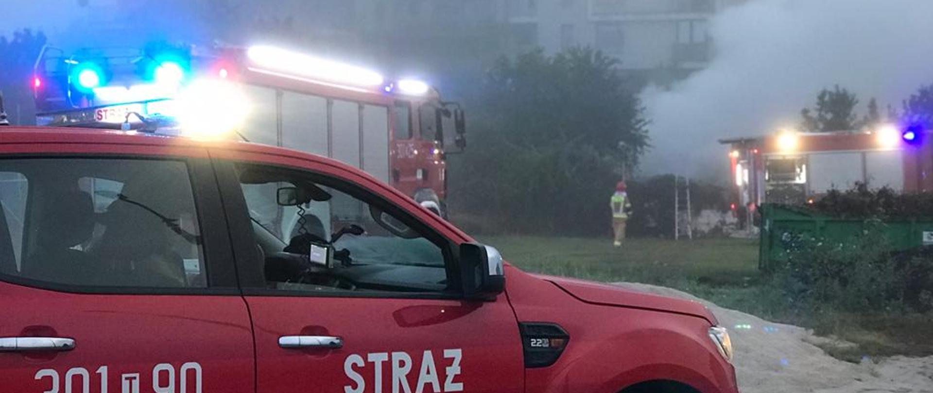 Na pierwszym planie stoi samochód pożarniczy a za nim drugi. Po prawej stronie widać unoszący się z pożaru dym a obok kolejny wóz strażacki.