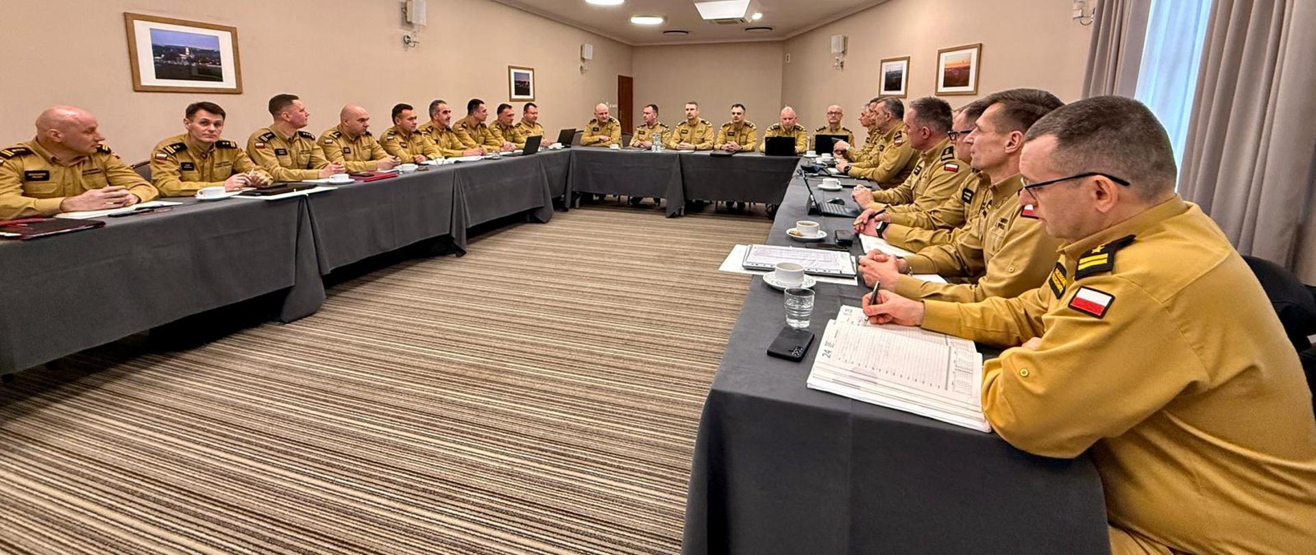 Strażacy podczas narady w mundurach służbowych siędzą przy stołach.