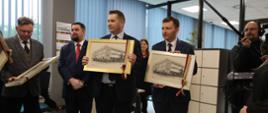 Minister Czarnek stoi i trzyma przedstawiający budynek, czarno-biały obrazek w ramkach, obok niego mężczyzna w garniturze z takim samym obrazkiem, za nimi kilka osób.