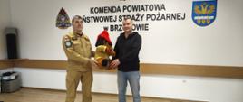 Kolorowa fotografia wykonana w pomieszczeniu świetlicy KP PSP w Brzozowie. Przedstawia strażaka PSP w umundurowaniu służbowym (piaskowym) podczas wręczania na licytację maskotki tzw. Żarka dla sztabu WOŚP w Warze. W tle czarny napis na ścianie Komenda Powiatowa Państwowej Straży Pożarnej w Brzozowie oraz logo PSP i powiatu brzozowskiego.