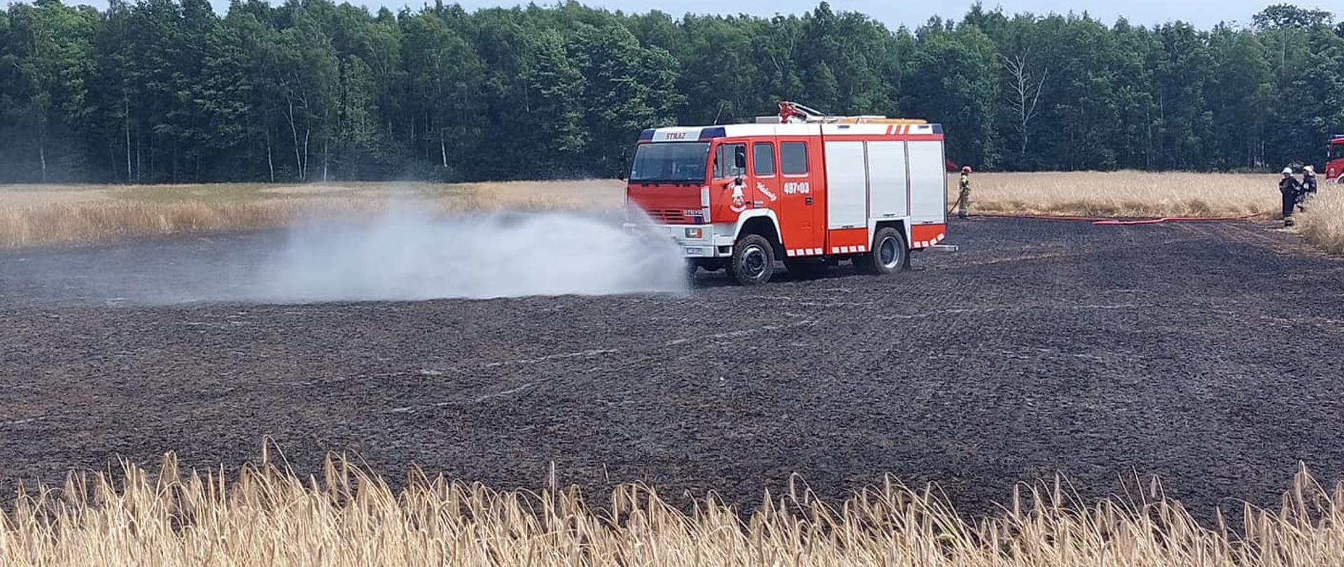 Na zdjęciu znajduje się wóz straży pożarnej na polu podający wodę ze zraszaczy, z tyłu widać strażaków i zboże na pniu