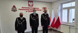 Na zdjęciu w sali konferencyjnej trzech druhów OSP w ubraniach galowych koloru granatowego bez czapek. W tle biała ściana na której znajduje się godło logo PSP i herb powiatu gorlickiego oraz trzy biało czerwone flagi.