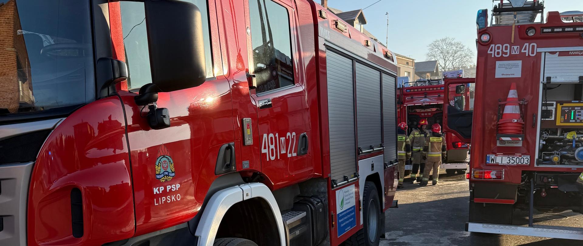 Pożar Lipsko ul. Czachowskiego pojazdy podczas działań. Zdjęcie przedstawia dwa czerwone wozy strażackie stojące na utwardzonym placu w słoneczny dzień. Po lewej stronie kadru widoczny jest przód i bok jednego z wozów, z numerem "481M22" na kabinie i logo KP PSP Lipsko. Po prawej stronie, nieco dalej, widać tył drugiego wozu z numerem "489M40".
W tle, pomiędzy pojazdami, widać grupę strażaków w ciemnych mundurach. Na horyzoncie, za budynkami, widoczne są gałęzie drzew i słupy. Niebo jest błękitne z delikatnymi smugami chmur.