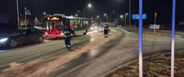 Strażacy z miotłami usuwają plamę oleju z drogi w tle autobus miejski oraz samochód osobowy.