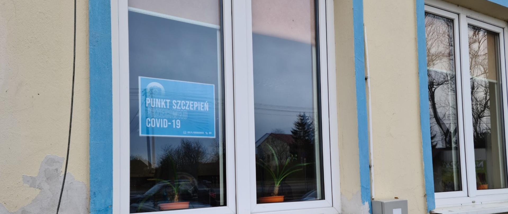 Fragment budynku - punktu szczepień. budynek w kolorze kości słoniowej, okna o ościeżnicach białych. W niebieskim kolorze obramowanie okien. Za szybą tabliczka z napisem - punkt szczepień przeciw cowid-19 