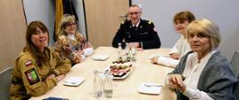 Pięć osób siedzi przy jasnym, prostokątnym stole w sali konferencyjnej. Po lewej stronie widać kobietę w beżowym mundurze strażackim, obok niej dwie kobiety w cywilnych strojach, natomiast po prawej stronie – mężczyznę w czarnym mundurze strażackim (komendant wojewódzki) oraz kolejną kobietę w stroju cywilnym. Na środku stołu stoi patera z pokrojonym ciastem oraz kilka butelek wody. W tle widoczne są jasne drzwi i fragment garderoby na wieszaku.