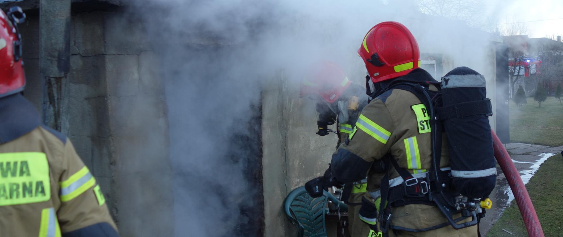 Strażacy w aparatach ochrony dróg oddechowych gaszą pożar budynku. W środku panuje duże zadymienie.