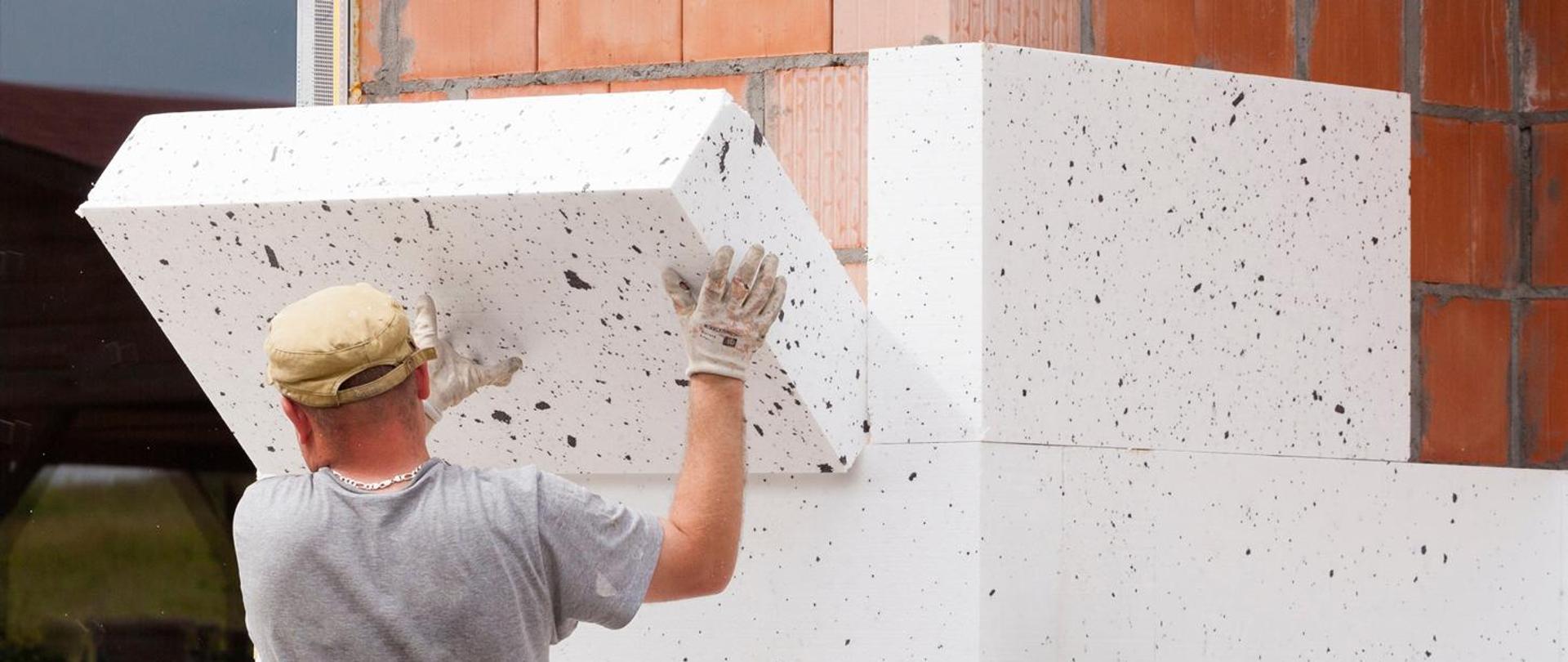 Mężczyzna w szarej koszulce, białych rękawicach oraz beżowej czapce układa na elewacji budynku biały styropian.