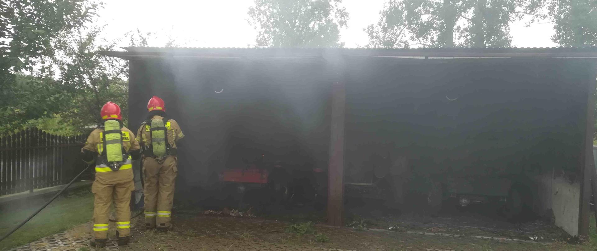 Zdjęcie zrobione w dzień. Na zdjęciu widać palący się garaż. Zdjęcie przedstawia dwóch strażaków ubranych w sprzęt ochrony układu oddechowego gaszących palący się garaż. 