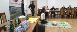 Kobieta ćwiczy zabiegi resuscytacyjne na fantomie noworodka. Strażacy prowadzą instruktarz. W tle osoby przyglądające się prelekcji.