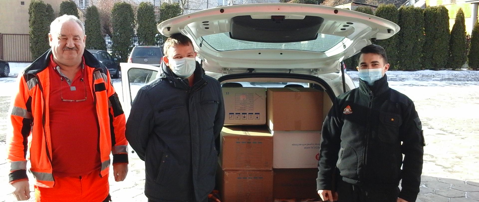 Zdjęcie przedstawia trzy osoby, tj. (od lewej) dwaj pracownicy Szpitala Ogólnego w Kolnie oraz strażak JRG Kolno. W tle pojazd z otwartą przestrzenią bagażową, w której znajdują się kartony z maseczkami.