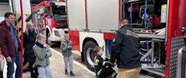 Na zdjęciu znajduje się wóz strażacki stojący na garażu, obok samochodu ustawione jest czworo dzieci oraz dwie osoby dorosłe, przy samochodzie stoi strażak w umundurowaniu służbowym.