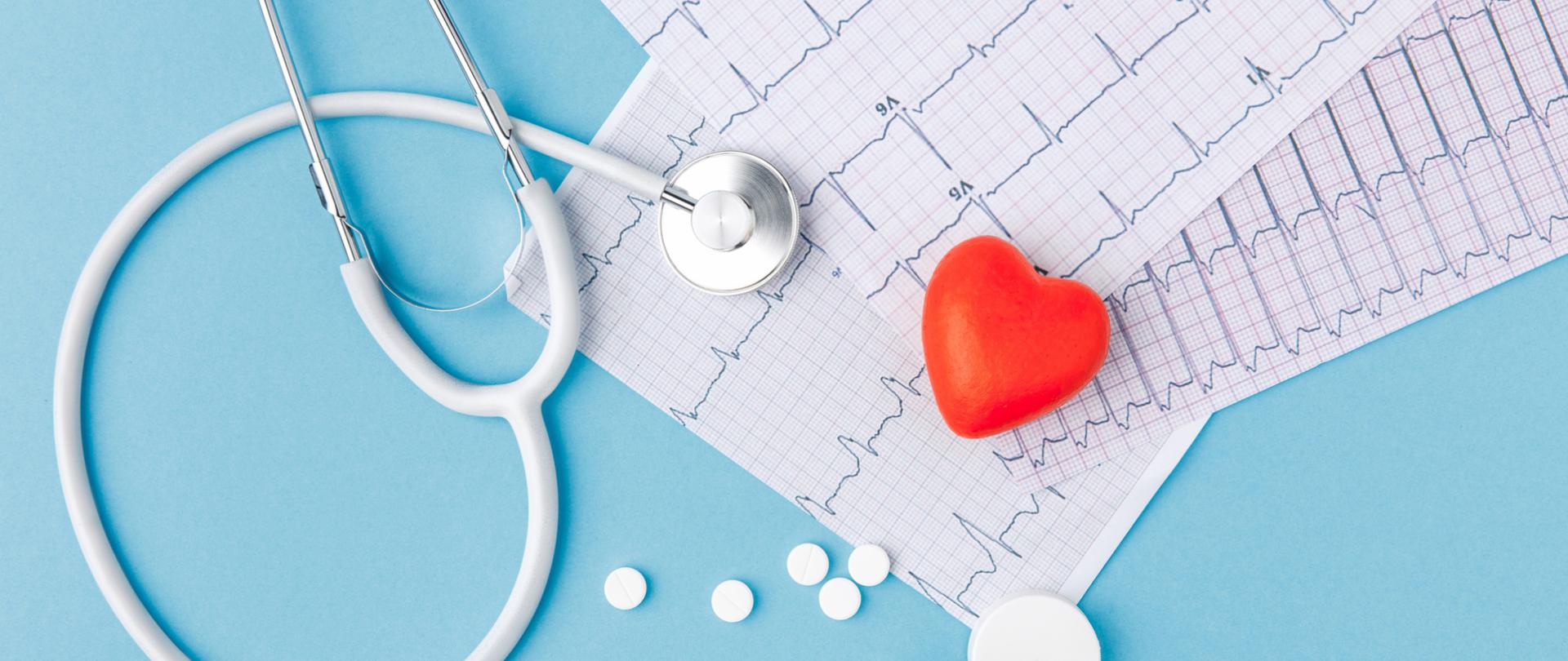 Na zdjęciu są wydruki ekg, stetoskop, tabletki oraz serce. Tło jest niebieskie.