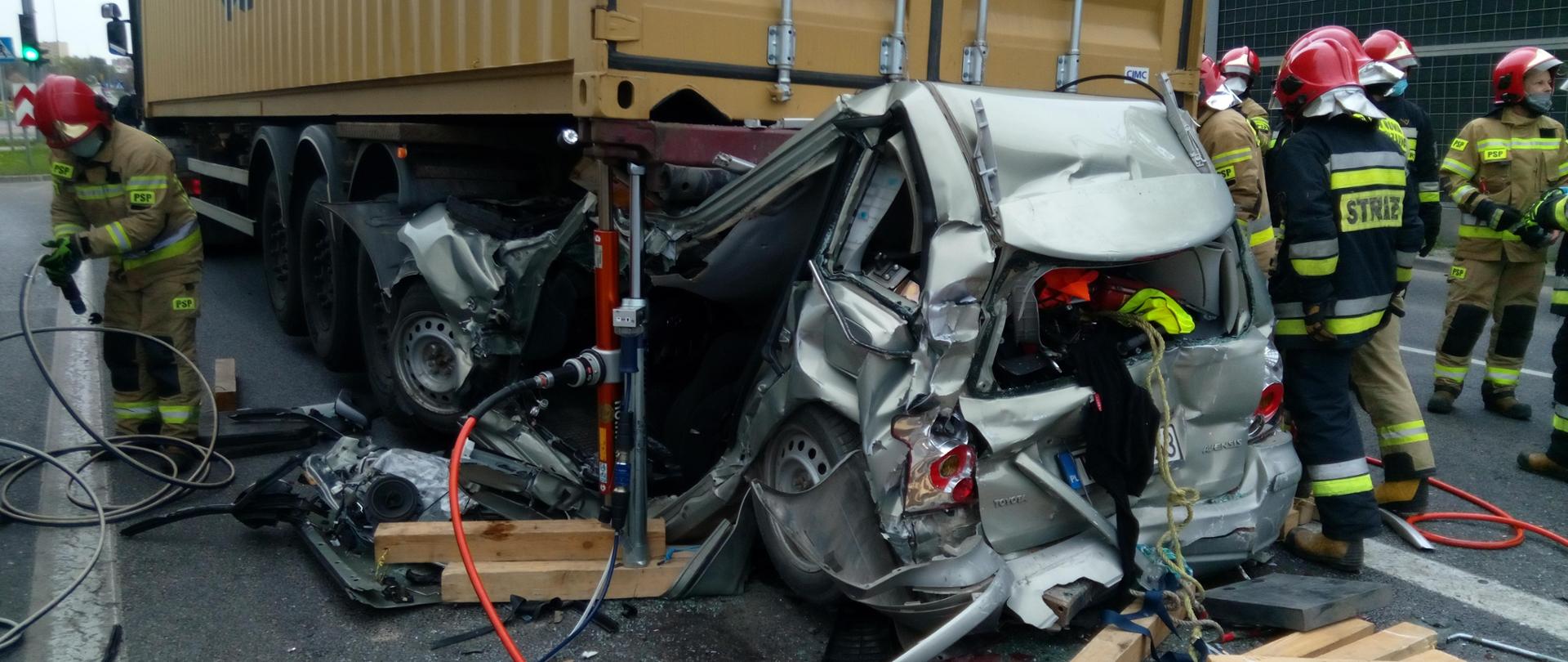 Zdjęcie przedstawia całkowicie zniszczony zamochód osobowy, który znajduje się z tyłu pod naczepą samochodu ciężarowego. Wokół pojazdów znajdują się strażacy, którzy wykonują swoje zadania.