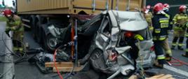Zdjęcie przedstawia całkowicie zniszczony zamochód osobowy, który znajduje się z tyłu pod naczepą samochodu ciężarowego. Wokół pojazdów znajdują się strażacy, którzy wykonują swoje zadania