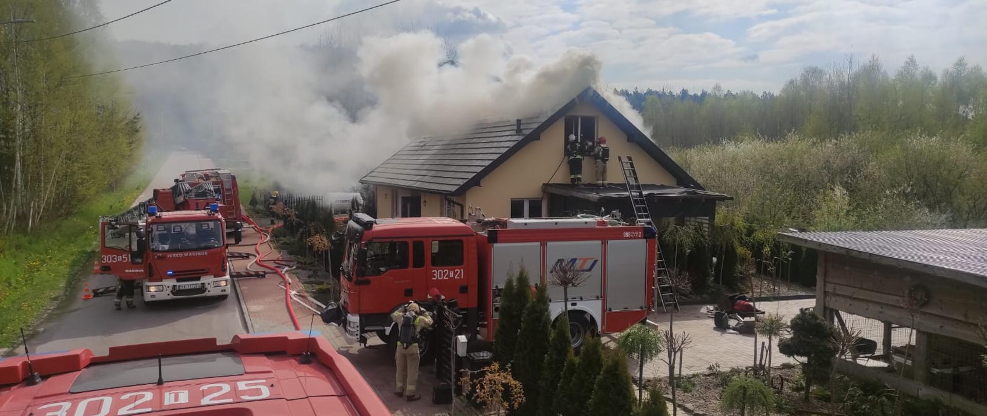 Zdjęcie przedstawia budynek w którym wybuchł pożar. Wokół domu stoją samochody strażackie. Z dachu unosi gęsta chmura dymu i pary wodnej.