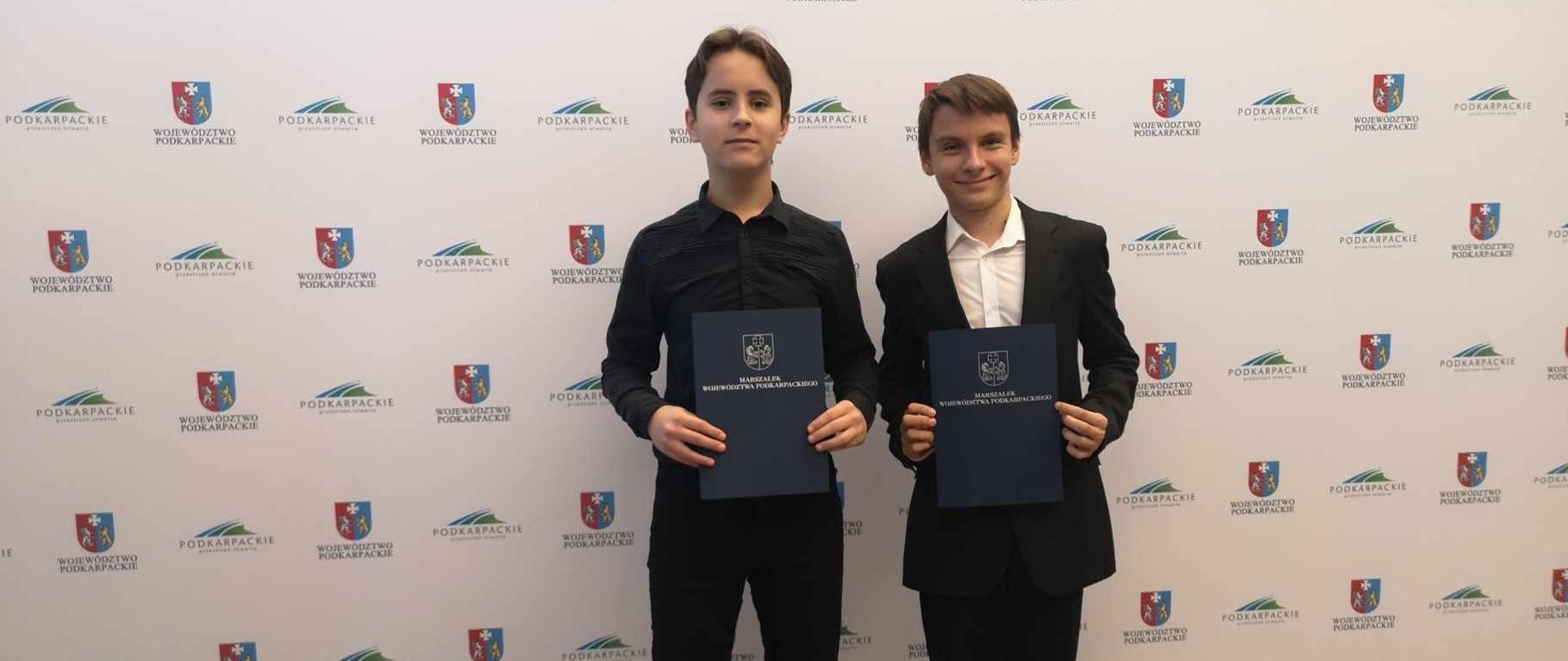 Dwoje uczniów w strojach galowych trzyma w rękach dokument potwierdzający przyznanie stypendium. W tle na białej ścianie logotypy Zarządu Województwa Podkarpackiego.